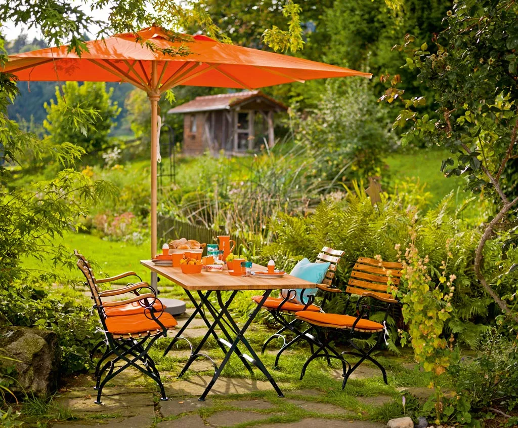 Essgruppe Classic von Weish in Orange auf einer Terrasse im Garten mit Holztisch und Stühlen, umgeben von üppigem Grün und Sonnenschirm.