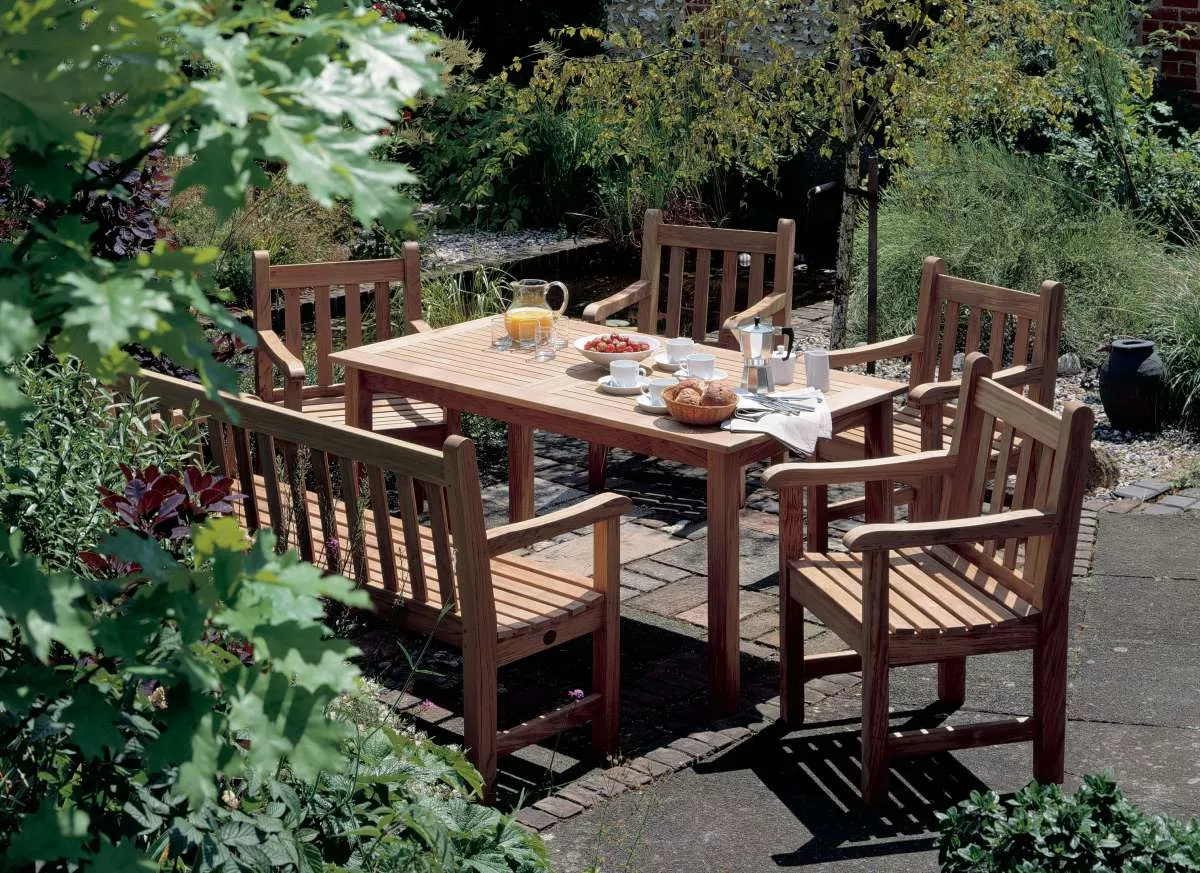 Gartenmöbel-Set von Barlow Tyrie aus Teakholz, bestehend aus einem 150 cm langen Tisch und sechs Stühlen, auf einer Terrasse im Grünen.