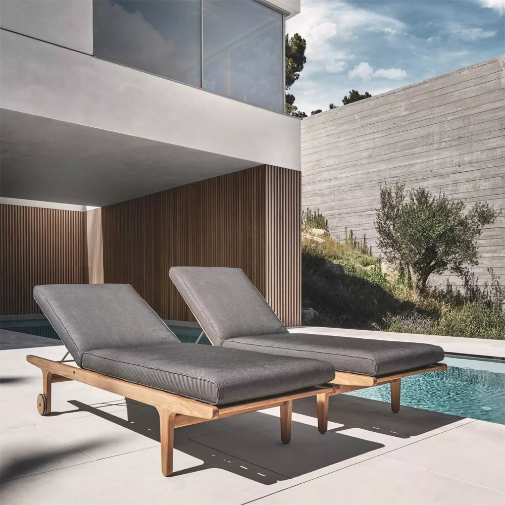 Zwei graue Gloster Bay Sonnenliegen aus Holz stehen nebeneinander auf einer Terrasse neben einem Pool, umgeben von moderner Architektur.