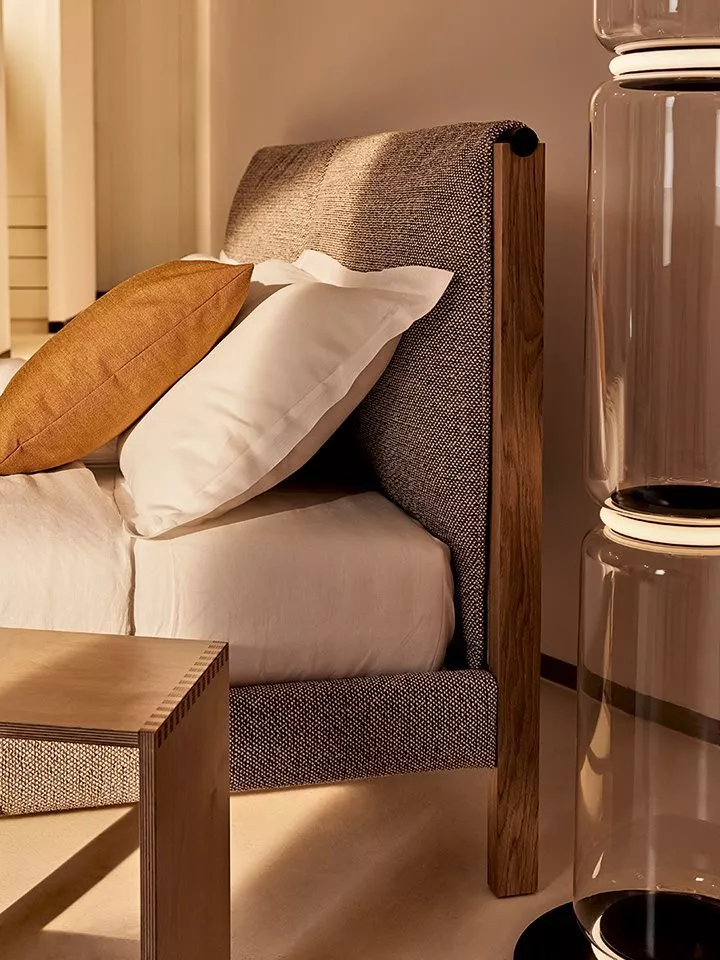 Das Bild zeigt das Zanotta Ricordi Bett mit Holzrahmen und grauem Stoffbezug in einem gemütlichen Schlafzimmer mit Kissen und Nachttisch.