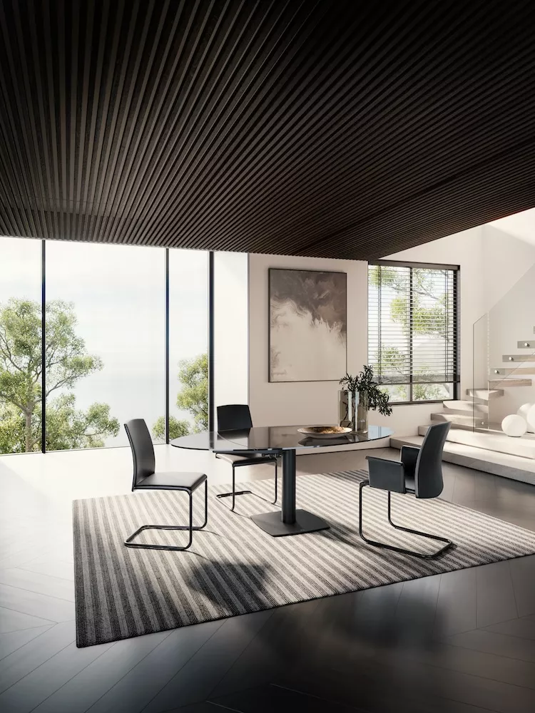 Ein moderner Essbereich mit einem Titan Draenert Tisch und vier schwarzen Stühlen in einem lichtdurchfluteten Raum mit großen Fenstern und Naturblick.