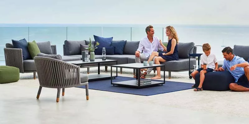 Cane-line Moments Loungegruppe in Grau auf einer Terrasse mit Meerblick, bestehend aus Sofas, Sesseln und Tischen, in entspannter Atmosphäre.