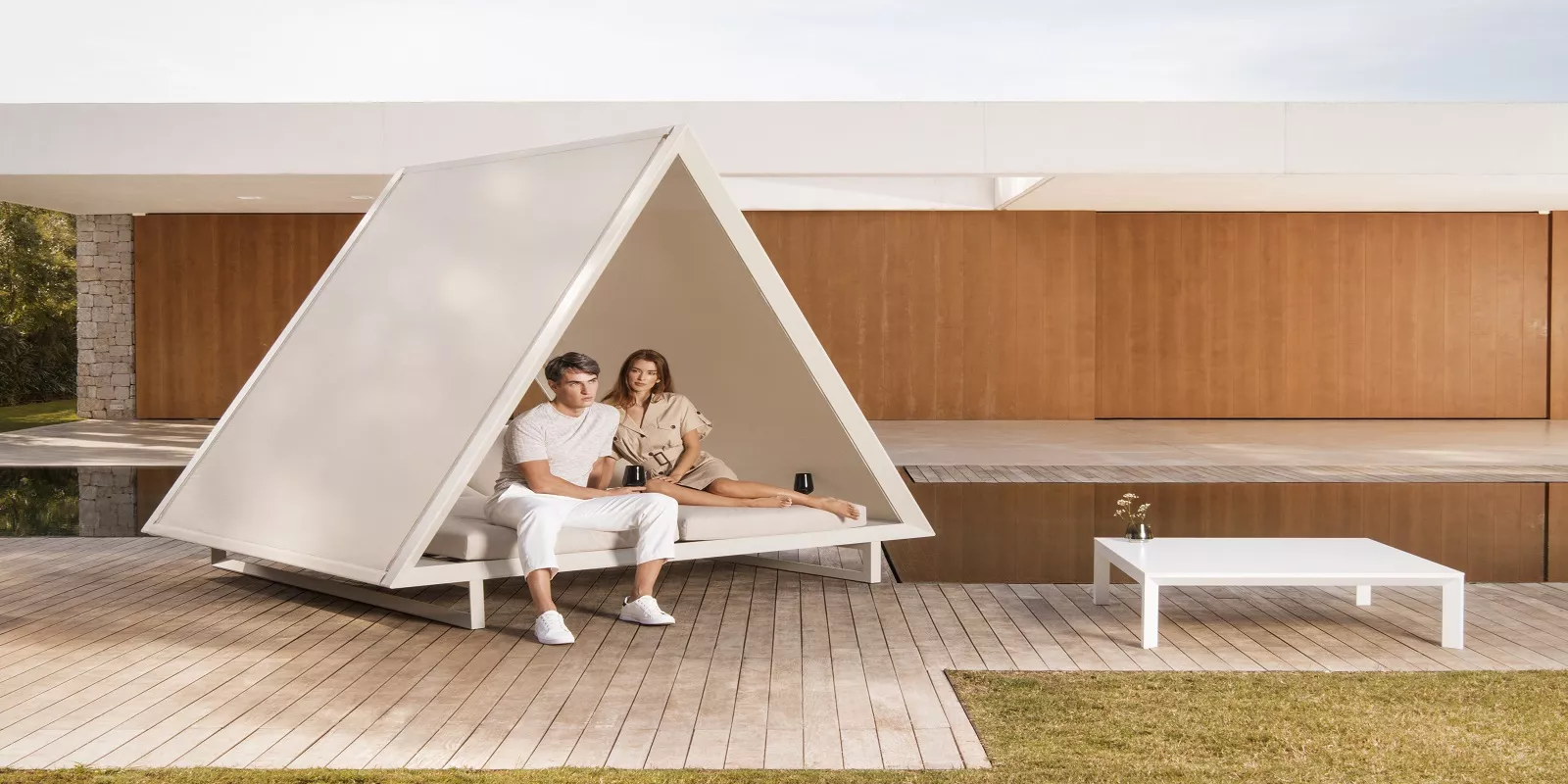 Ein Paar entspannt sich in einem weißen Vondom Vineyard Pavillon von Ramon Esteve auf einer Holzterrasse vor einem modernen Gebäude.
