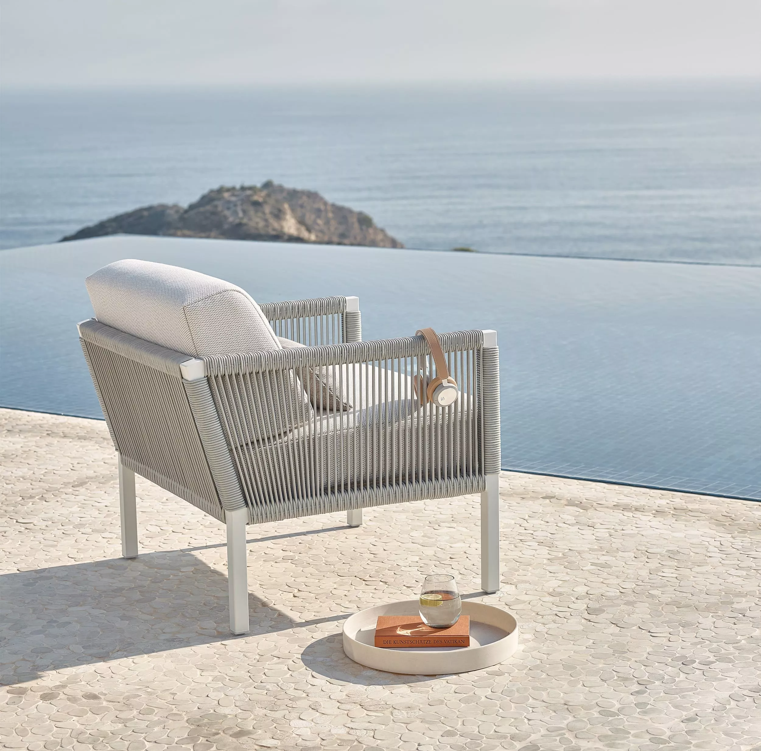 Solpuri Club A Sessel steht auf einer Terrasse mit Blick auf das Meer und einen Infinity-Pool im Hintergrund Moodbild mit Kopfhörern und Getränk.