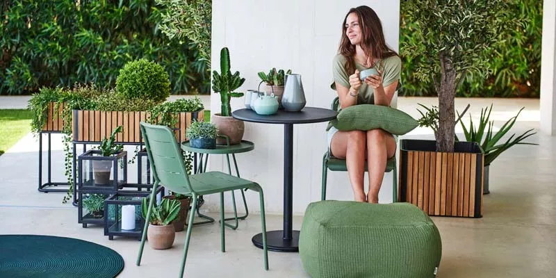 Frau sitzt entspannt an einem Cane-line Combine Essgruppe in Grün auf einer Terrasse mit Pflanzen im Hintergrund.