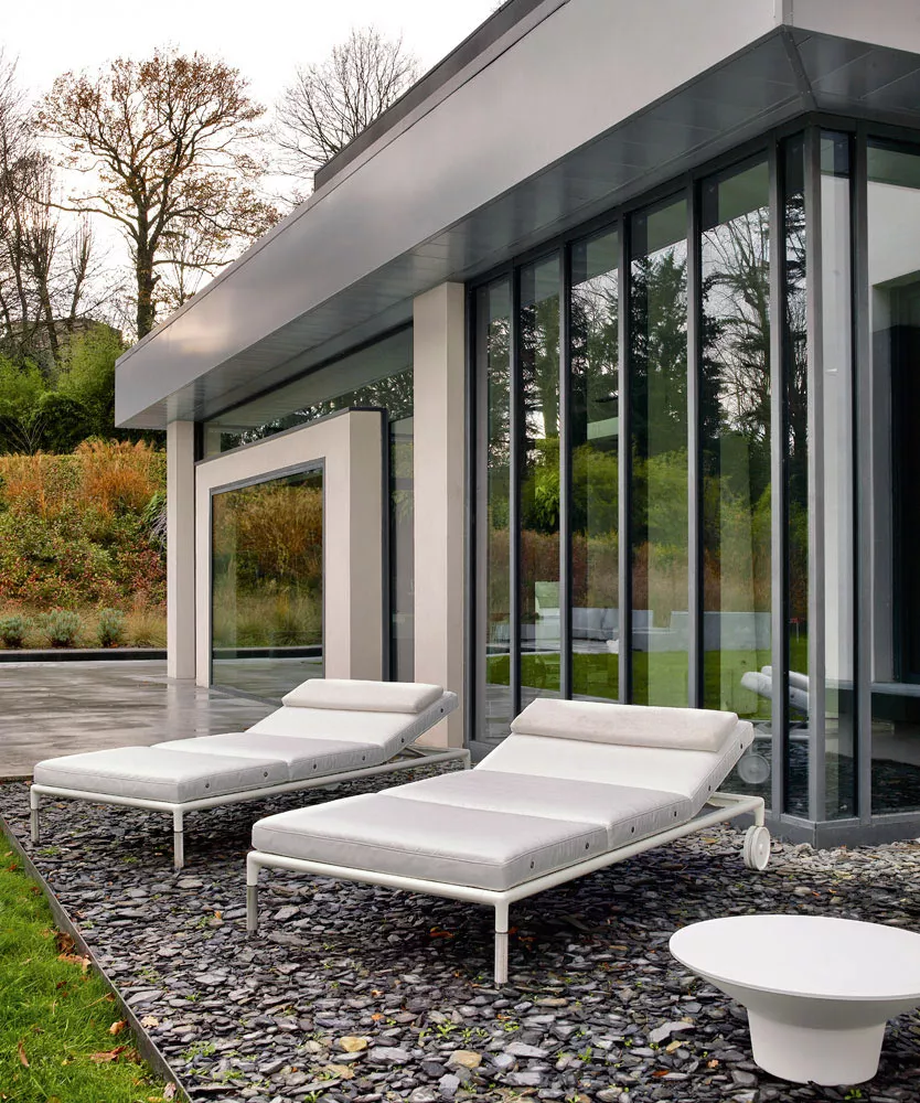 B&B Italia Springtime Outdoor Beistelltisch 43 cm steht neben zwei weißen Sonnenliegen vor einem modernen Haus mit großen Glasfenstern im Garten.