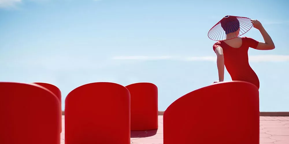 Frau in rotem Kleid und Hut sitzt auf rotem Diabla Lipstick Stuhl, vor blauem Himmel, moderne Outdoor-Szene.