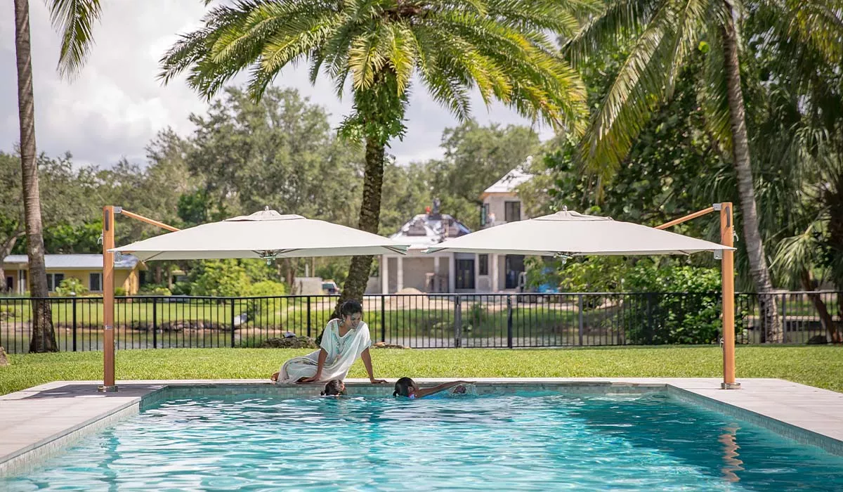 Zwei Bay Master Swim Sonnenschirme in Weiß spenden Schatten am Pool, umgeben von Palmen und einem luxuriösen Garten.