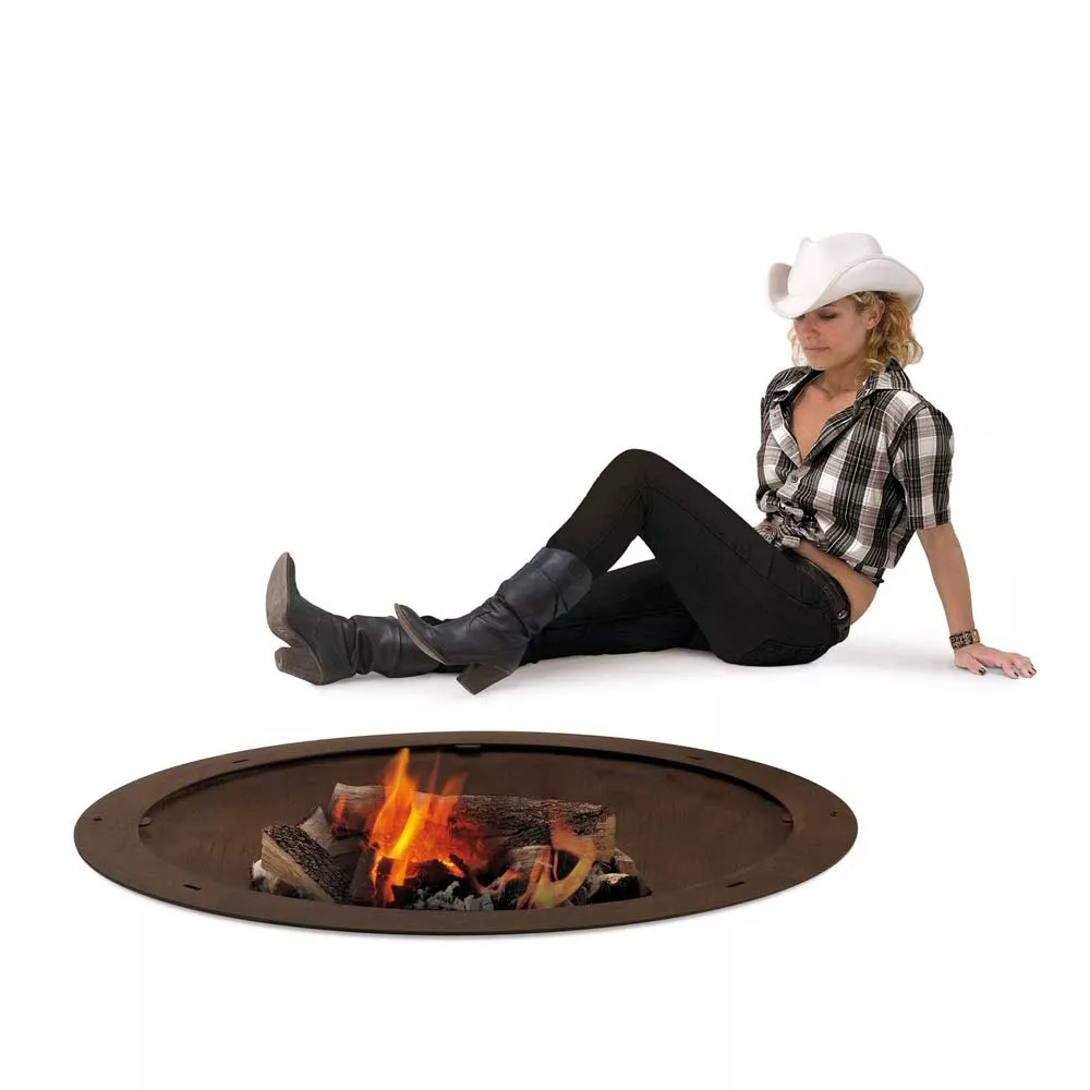 Eine Frau sitzt entspannt neben der AK47 Hole Feuerstelle aus oxidiertem Stahl und genießt das Feuer in einer gemütlichen Atmosphäre.