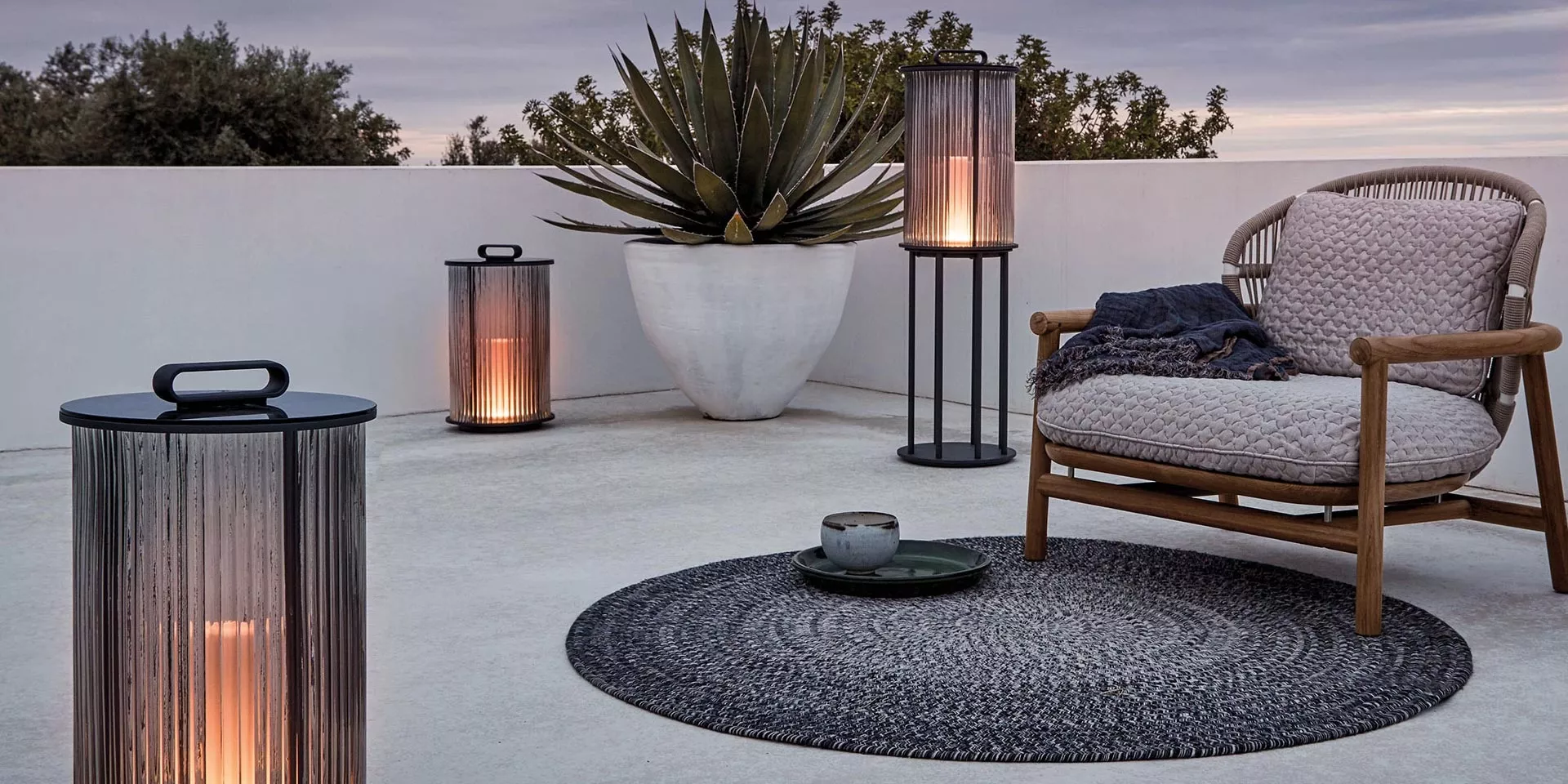 Gloster Outdoor-Leuchte in Schwarz auf einer Terrasse bei Dämmerung, umgeben von Pflanzen und Möbeln, stimmungsvolle Beleuchtung.