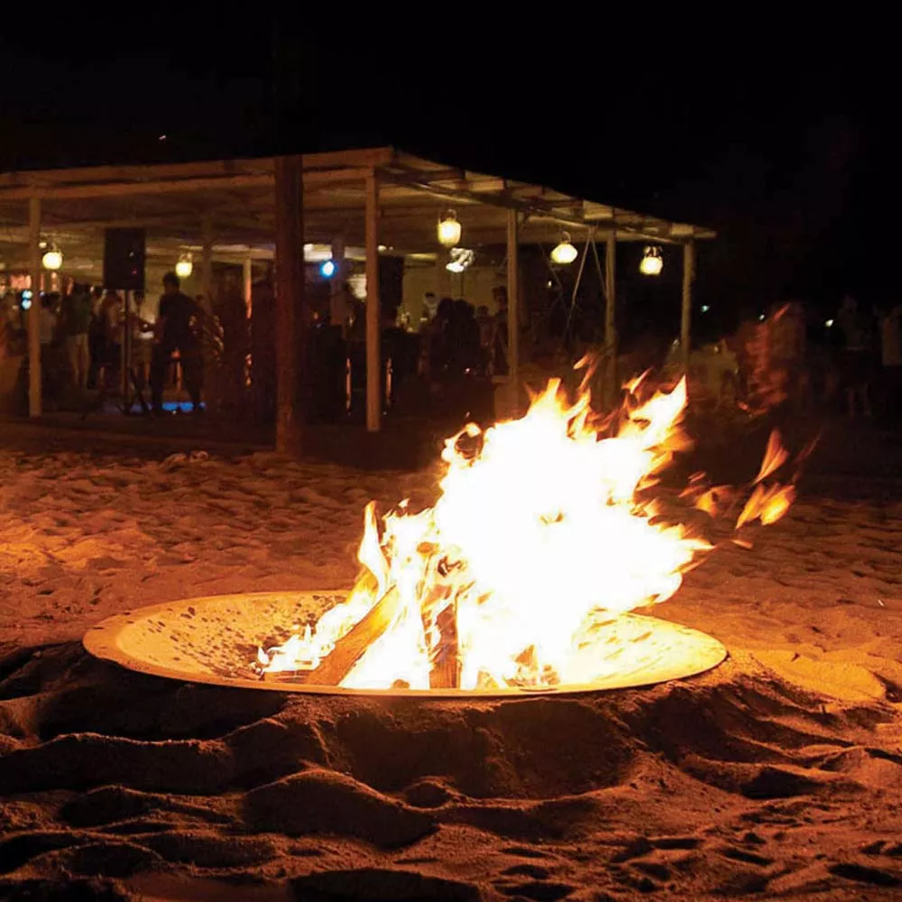 AK47 Hole Outdoor Feuerstelle brennend am Strand bei Nacht mit Menschen im Hintergrund die sich unter einem beleuchteten Pavillon versammeln
