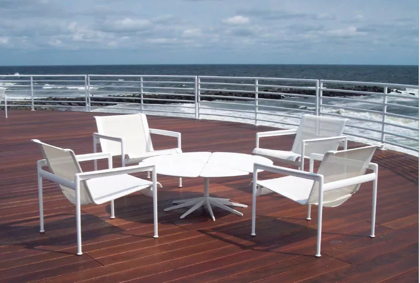 Vier weiße Knoll Studio Petal Stühle und ein passender Tisch stehen auf einer Holzterrasse mit Blick auf das Meer unter bewölktem Himmel