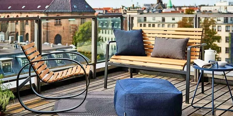 Cane-line Parc Lounge-Set in Holzoptik auf einer städtischen Dachterrasse mit blauem Hocker und Kissen, umgeben von Glasgeländer.