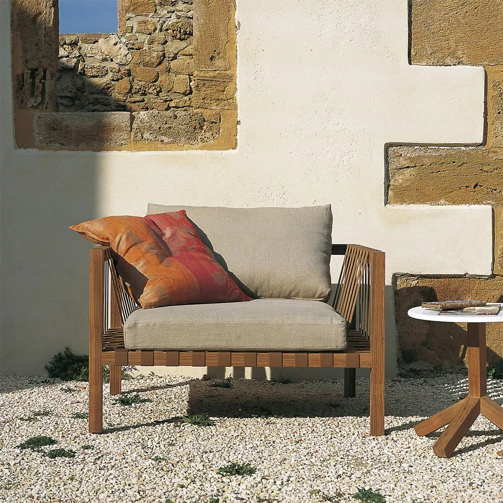 Roda Mistral Loungesessel 101 aus Teakholz in Naturfarbe auf Kiesboden vor einer Steinmauer mit blauem Himmel im Hintergrund.