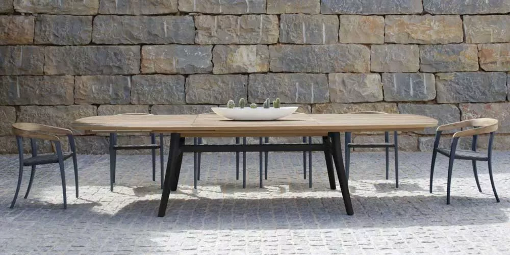 Royal Botania Zidiz Essgruppe in Naturholz und Schwarz auf gepflastertem Boden vor einer Steinmauer, stimmungsvolles Outdoor-Setting.
