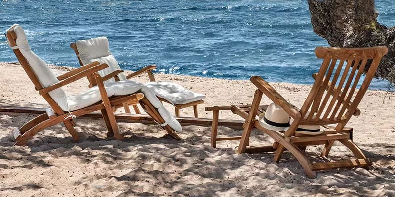 Zwei Ethimo Cruise Outdoor-Liegestühle aus Holz mit weißen Polstern stehen am Sandstrand vor dem Meer.