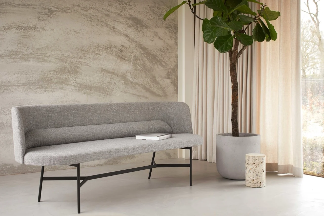 Graue HARRI Bank von more in modernem Wohnzimmer mit Betonwand und großem Fenster, neben einem hohen Pflanzentopf und einem kleinen Beistelltisch.