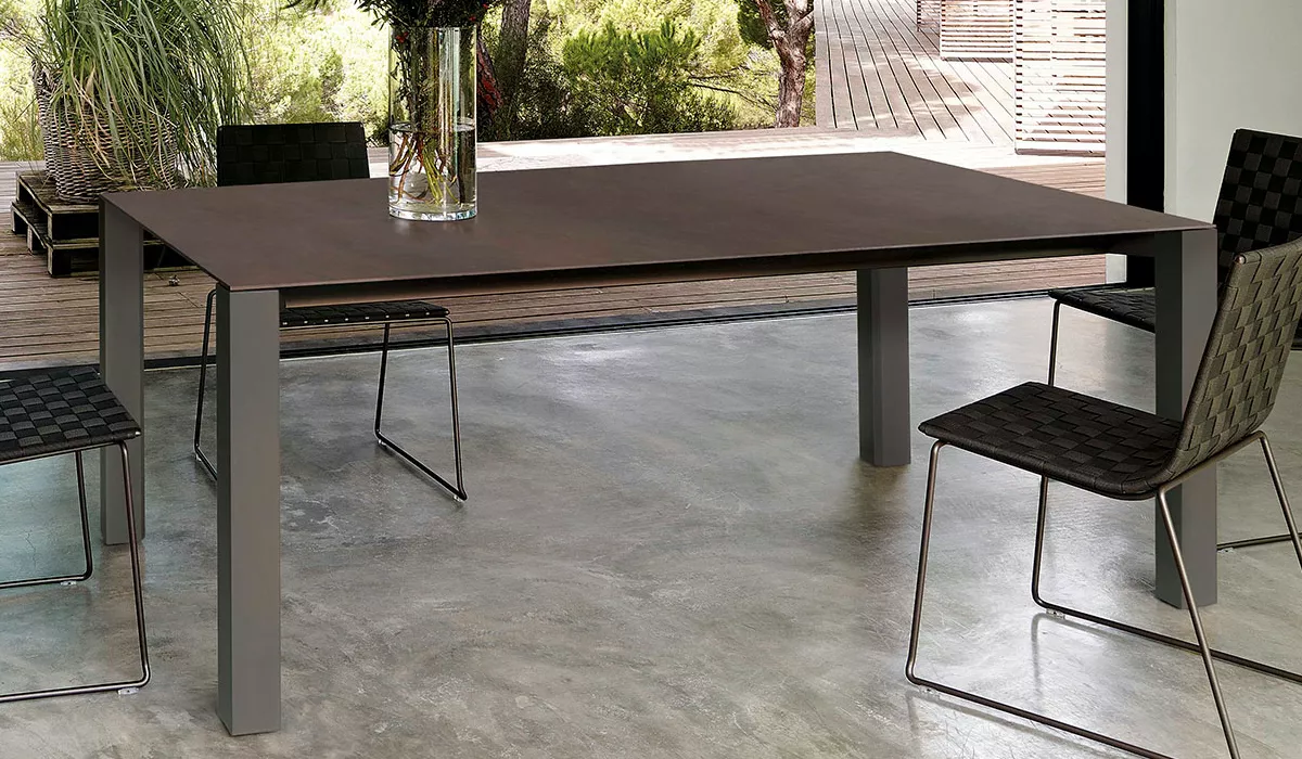 Brauner Andreu World Tisch in modernem Raum mit Betonboden und Glasvase, umgeben von geflochtenen Stühlen, Blick auf Terrasse.