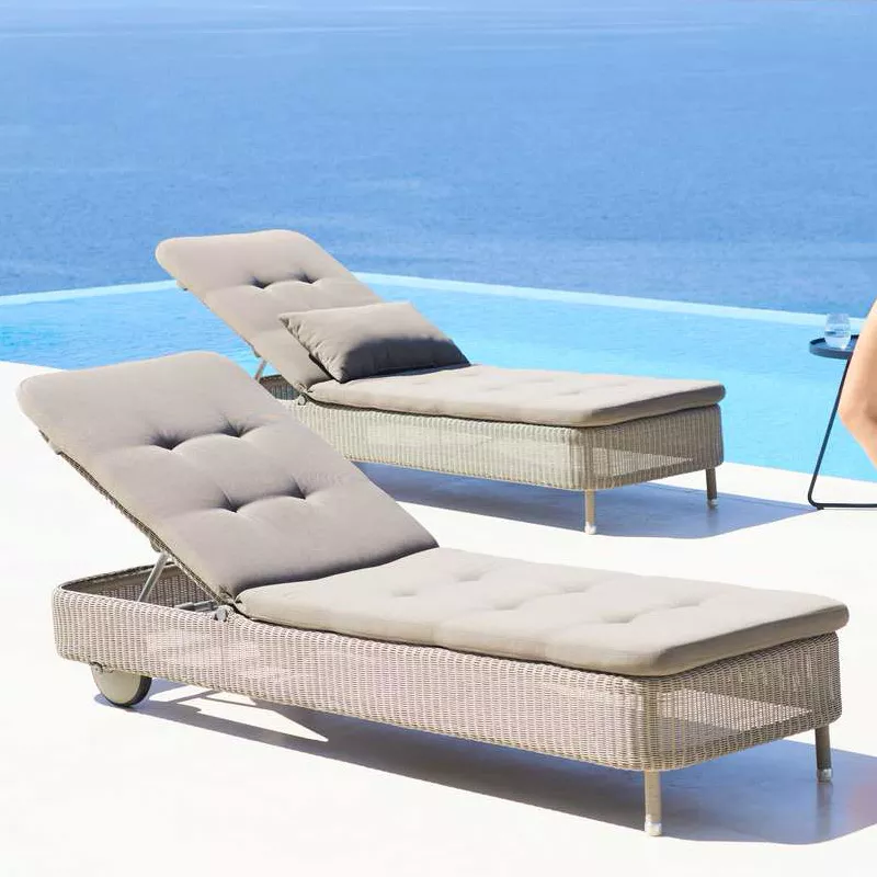 Zwei Cane-line Presley Sonnenliegen in Beige auf einer Terrasse mit Meerblick, entspannte Atmosphäre, seitliche Perspektive.