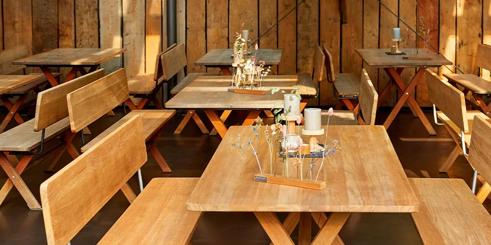 Weishäupl Cross Holztische und Bänke in natürlichem Holzton in einem rustikalen, holzverkleideten Raum mit dekorativen Kerzen auf den Tischen.