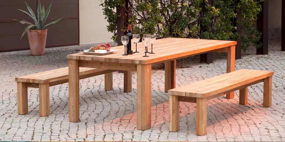 Barlow Tyrie Titan Esstisch-Set aus hellem Holz mit zwei Bänken auf gepflastertem Hof, dekoriert mit Kerzen und Wein.