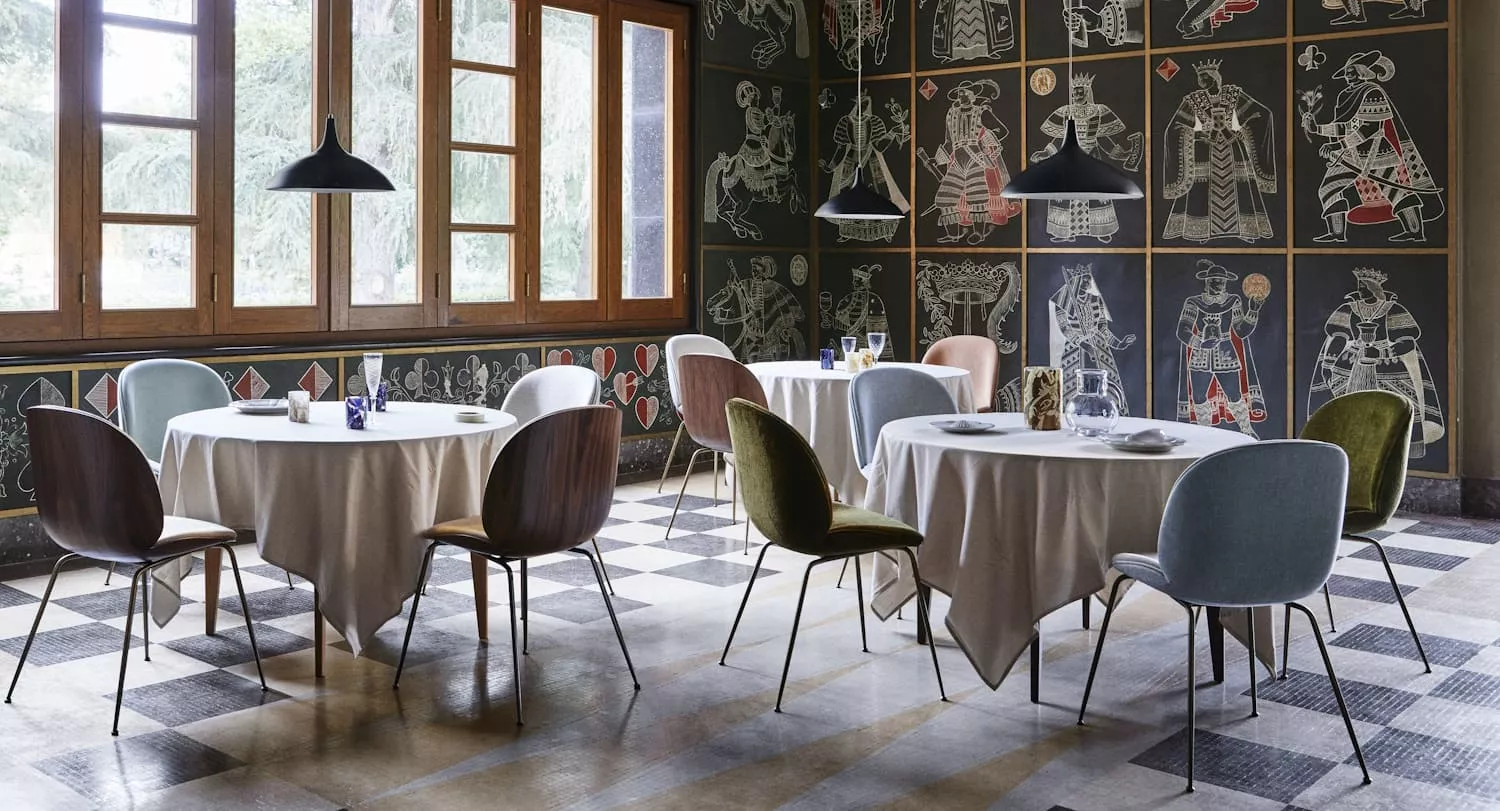 Ein stilvoll eingerichtetes Restaurant mit Gubi Stühlen und runden Tischen in einem Raum mit großen Fenstern und kunstvoll bemalten Wänden.