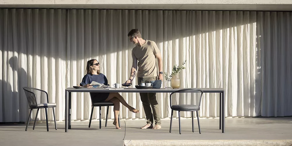 Ein Paar sitzt an einem modernen, grauen Todus Alca Tisch im Freien, umgeben von minimalistischer Architektur und sanftem Licht.