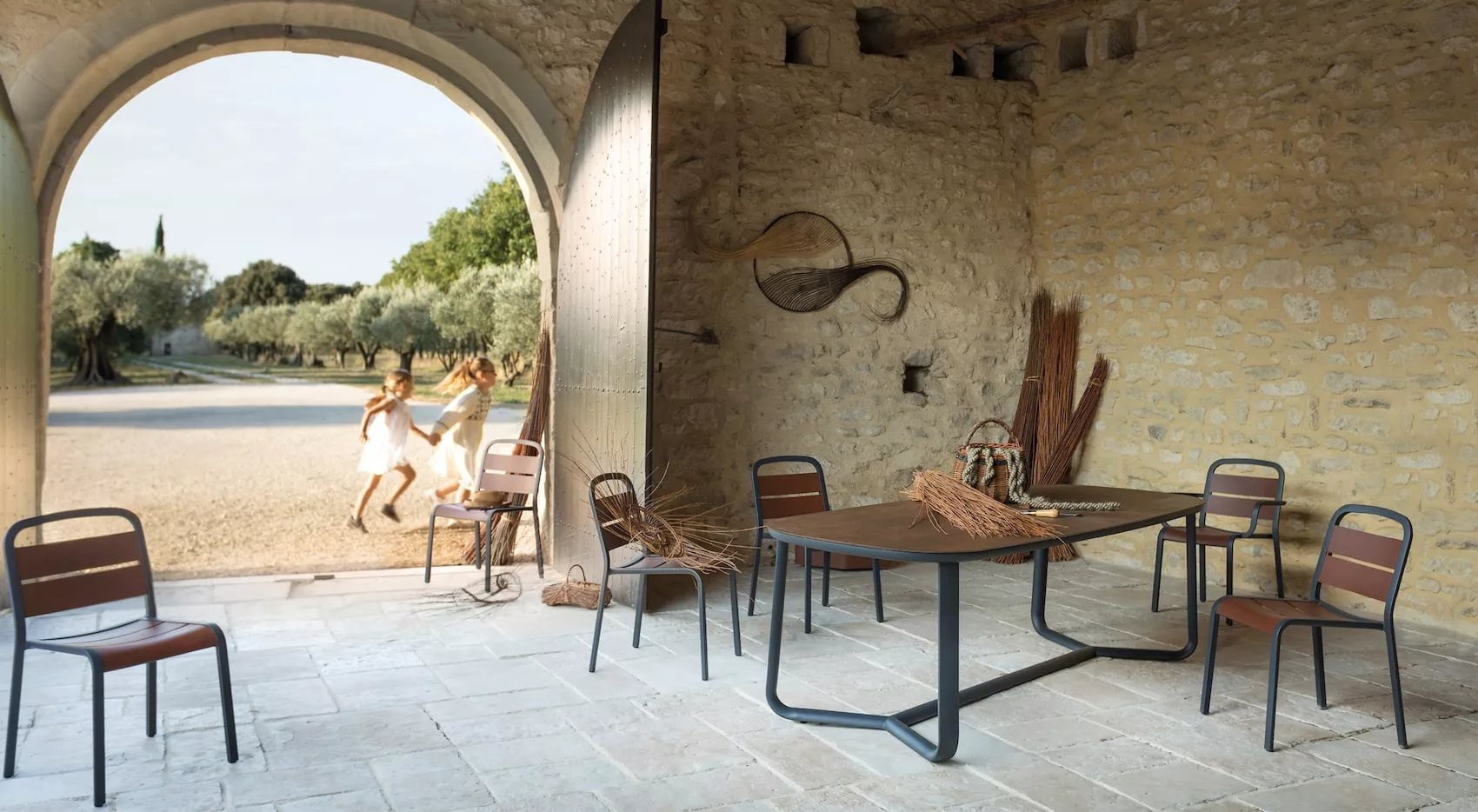 EGO Paris Marumi Esstisch 240 x 98 cm mit Teak-Tischplatte in rustikalem Innenhof, umgeben von Steinwänden und offenem Torbogen.