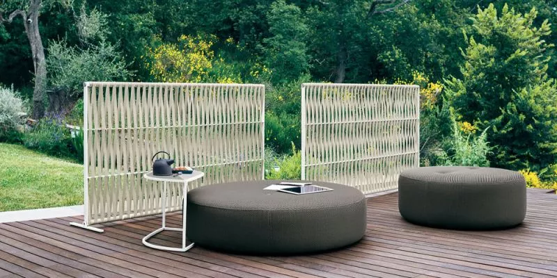 Zwei Roda Wing Paravents in Weiß auf einer Holzterrasse mit grüner Gartenkulisse, daneben runde, graue Sitzkissen und ein kleiner Tisch.