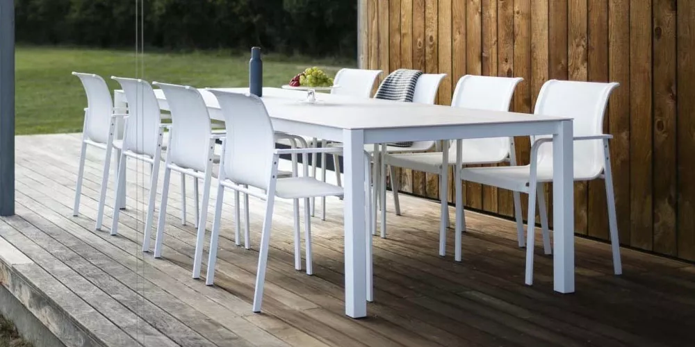 Todus Puro Tisch in Weiß auf einer Holzterrasse, umgeben von acht passenden Stühlen, mit Blick auf einen grünen Garten.