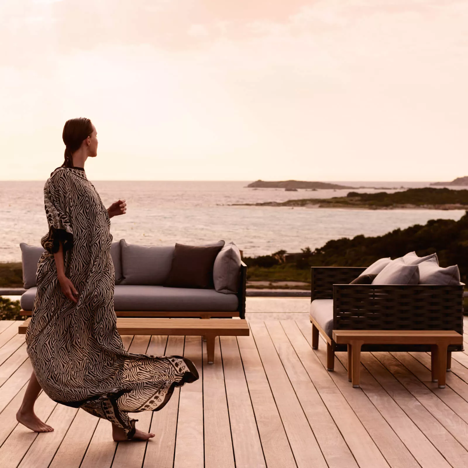 Eine Frau in einem langen Kleid geht an der Dedon Sealine Lounge vorbei auf einer Terrasse mit Meerblick im Hintergrund.