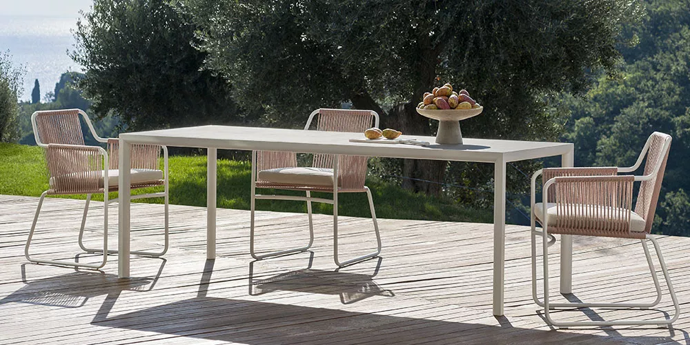 Roda Kollektion Plain Air, weißer Tisch mit vier Stühlen auf Holzterrasse, Hintergrund mit Bäumen und Meerblick, stimmungsvolles Bild.