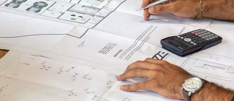 Ein Mann plant mit einem Stift auf einem technischen Bauplan, daneben ein schwarzer Taschenrechner auf einem Holztisch.