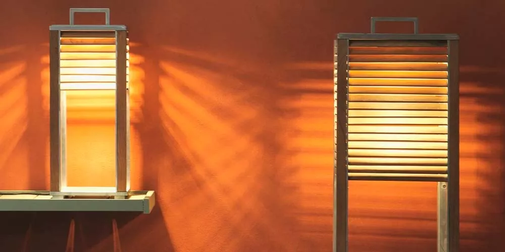 Zwei Ethimo Ginger Outdoor-Leuchten in warmem Licht vor einer orangefarbenen Wand, eine auf einem Regal, die andere auf dem Boden.