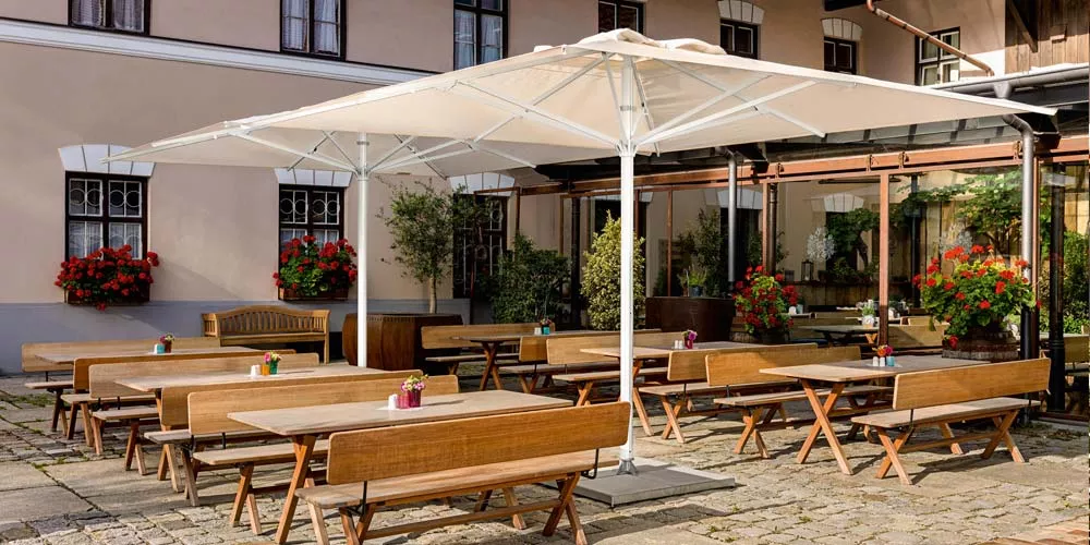 Weißer Weishäupl Teleskopschirm über Holzbänken in einem sonnigen Innenhof mit blühenden roten Geranien im Hintergrund.