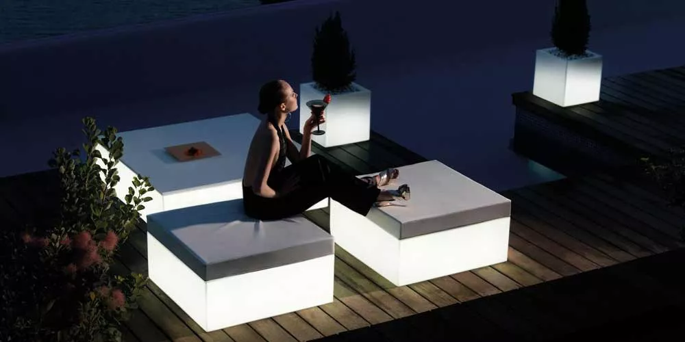 Frau sitzt abends auf beleuchteten, weißen Vondom Jut Chill Lounge-Gartenmöbeln auf einer Holzterrasse mit Pflanzen im Hintergrund.