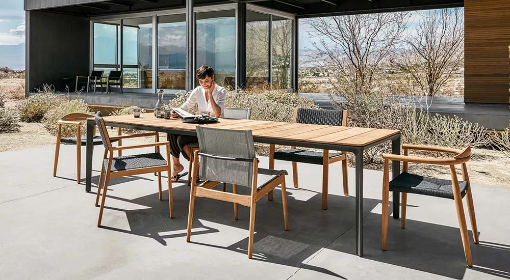 Frau sitzt an einem Gloster Carver Gartenmöbel Essgruppe aus Holz und Metall auf einer Terrasse mit modernem Haus im Hintergrund.