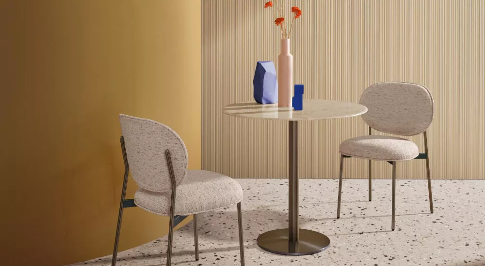 Mittelfuß-Tisch von Pedrali in Beige, umgeben von zwei Stühlen, auf einem Terrazzo-Boden vor einer gestreiften Wand, Stimmungsbild.