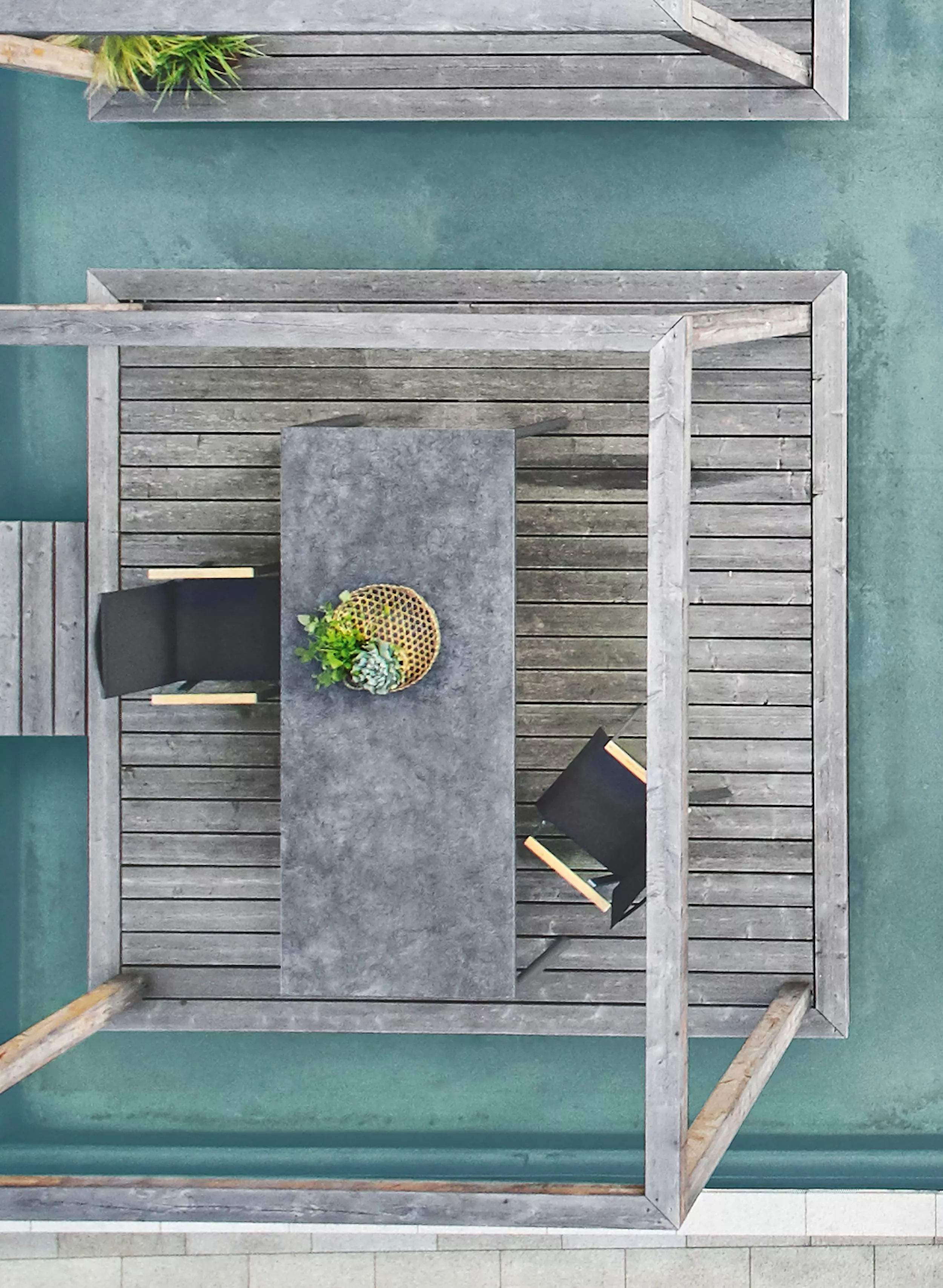 Zwei Solpuri Soul Stühle und ein Tisch mit einer Pflanze darauf stehen auf einer Holzterrasse über einem türkisfarbenen Pool in einer ruhigen Umgebung.