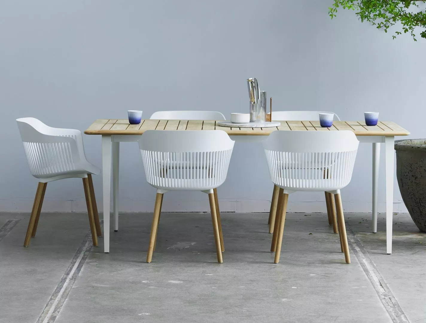 Dedon Aiir Essgruppe mit vier weißen Stühlen und einem Holztisch auf grauem Boden in einem minimalistischen Raum mit einer Pflanze im Hintergrund