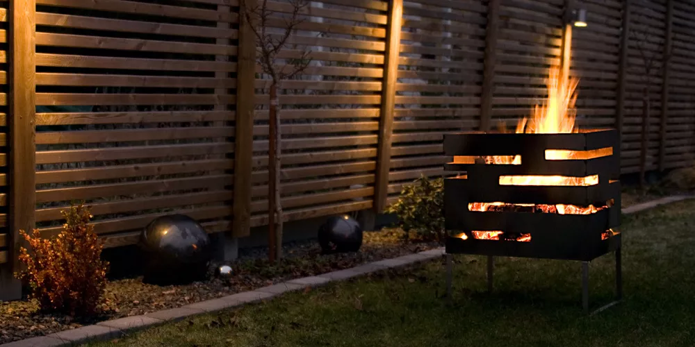 Roeshults Feuerkorb in Schwarz, stimmungsvoll beleuchtet im Garten vor einem Holzzaun, ideal für gemütliche Abende im Freien.