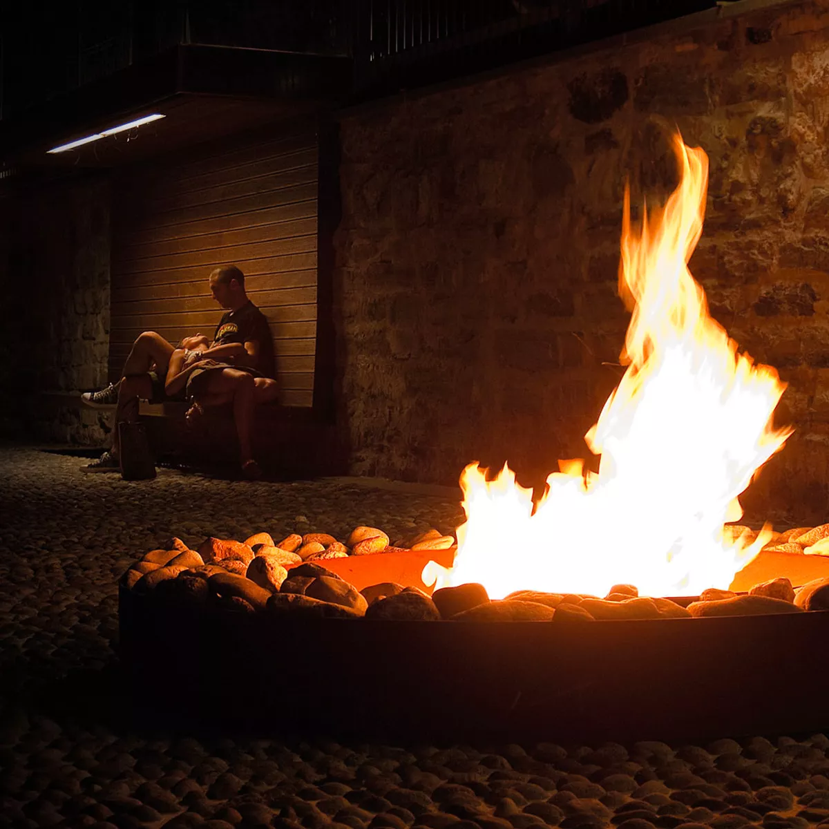 Ein Mann sitzt auf einer Bank und liest ein Buch während ein großes Feuer in der AK47 Zen Feuerstelle aus oxidiertem Stahl brennt.