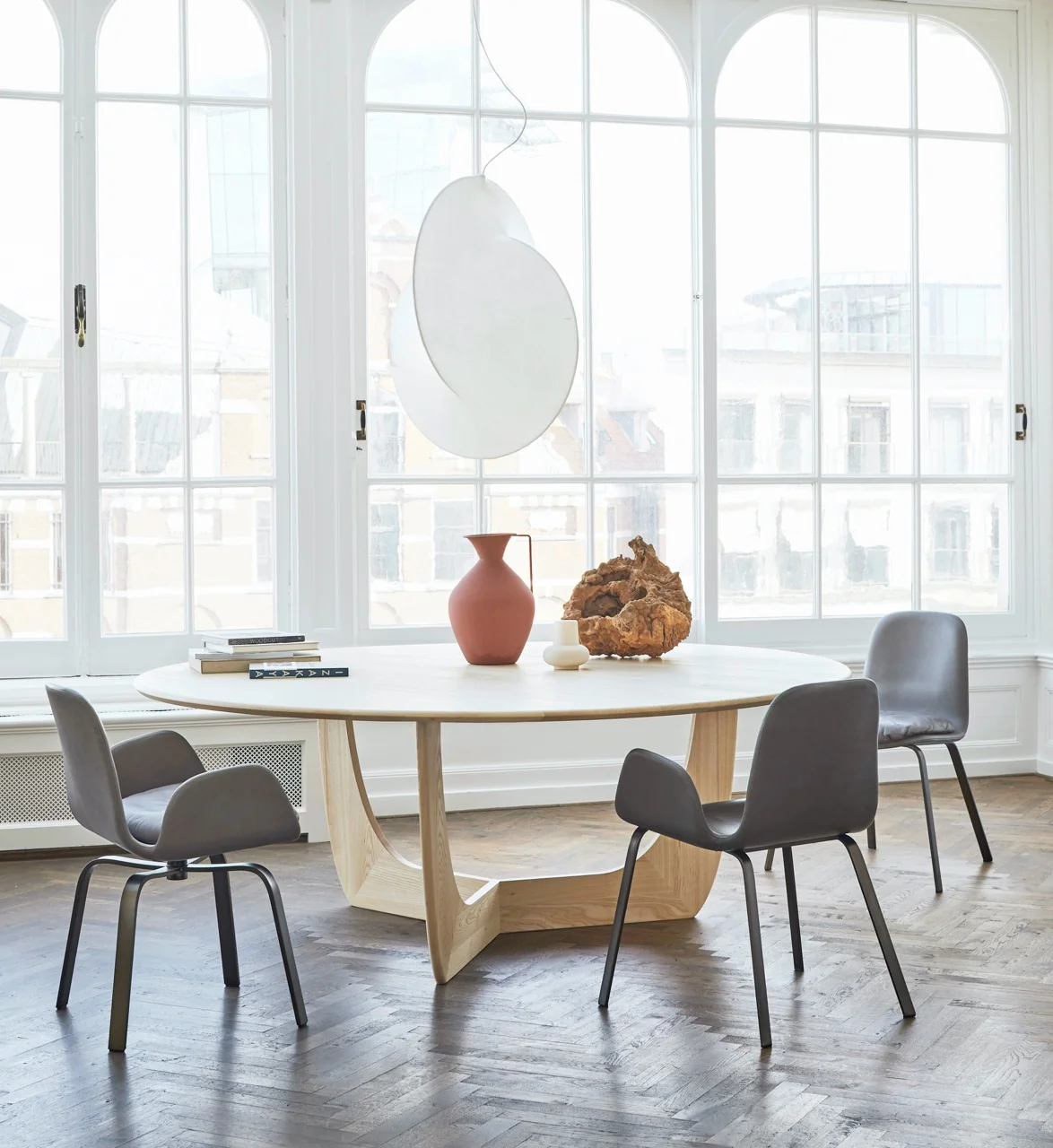 Runder CALA Tisch von more in hellem Holz mit grauen Stühlen in einem hellen Raum mit großen Fenstern und Parkettboden.
