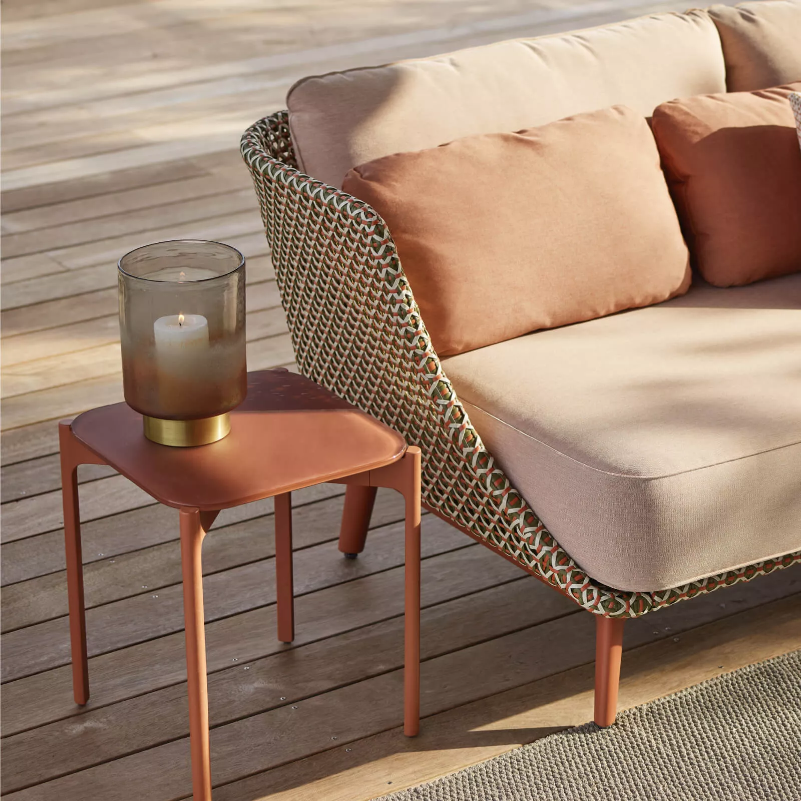 Dedon Mbarq 3er Sofa mit beigen Kissen und einem kleinen Beistelltisch mit Kerze auf einer Holzterrasse bei Tageslicht