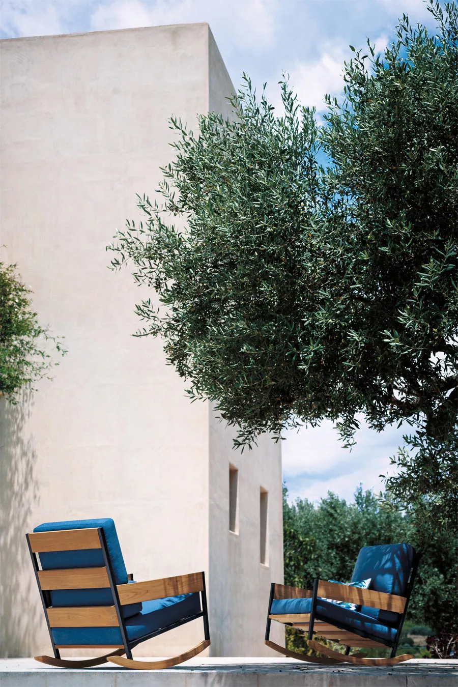 Zwei Roda Nap Schaukelsessel in Blau und Holzoptik stehen auf einer Terrasse vor einem modernen, beigen Gebäude mit Olivenbaum.