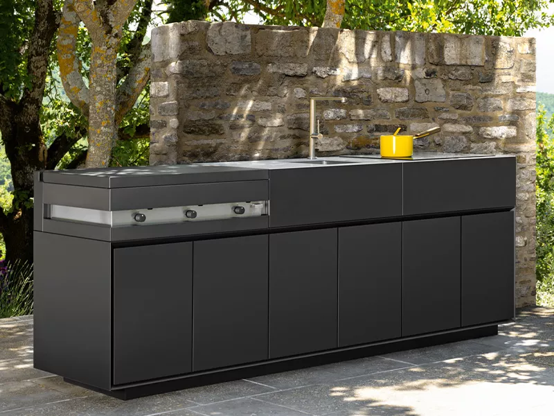 Talenti Outdoor Küche Tikal in Anthrazit vor einer Steinmauer im Garten, mit integriertem Grill und Spüle, stimmungsvolle Umgebung.