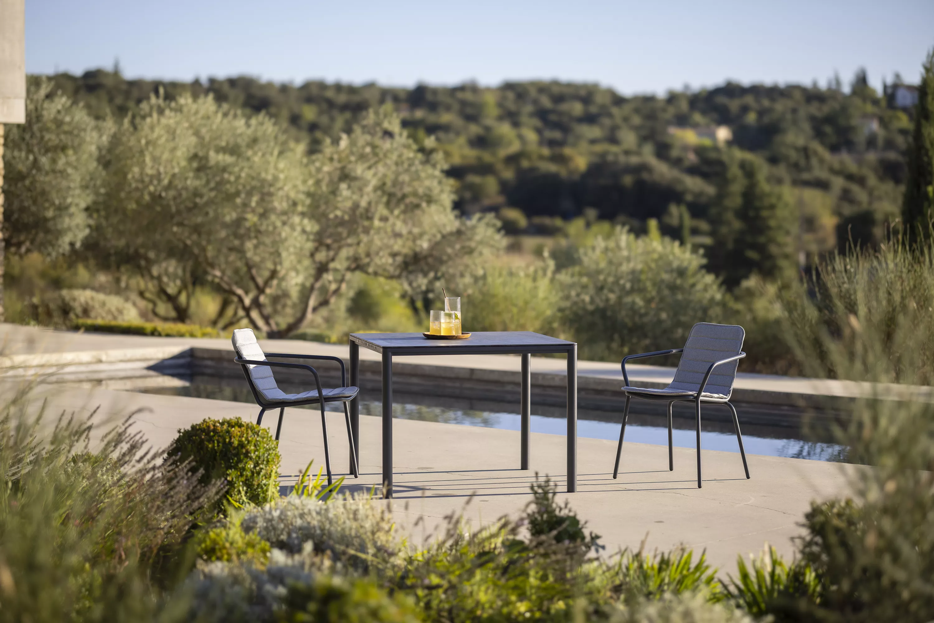 Todus Alca Esstisch in Schwarz mit HPL-Tischplatte, umgeben von zwei Stühlen, auf einer Terrasse mit Blick auf einen Garten.