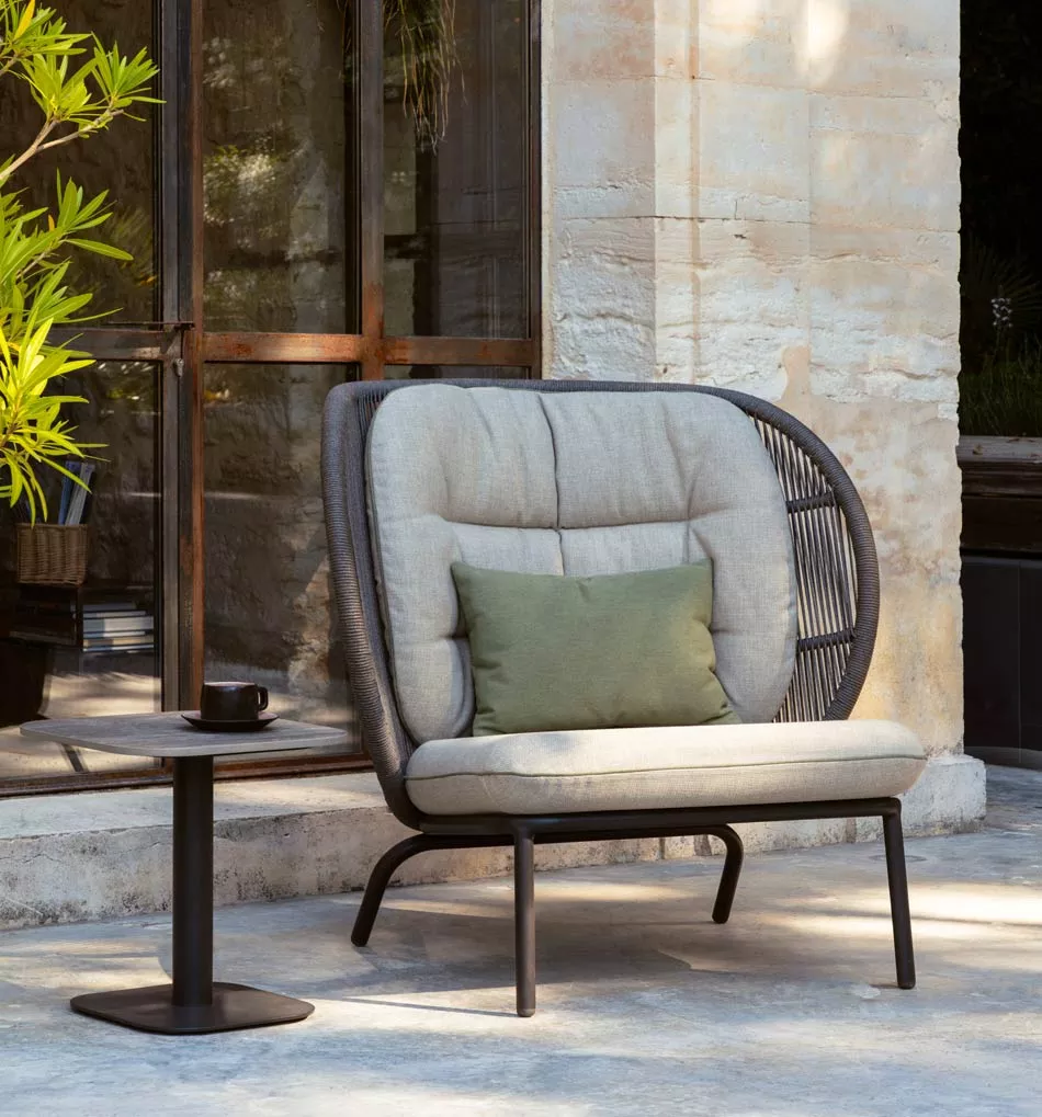 Vincent Sheppard Kodo Cocoon Outdoor Loungesessel in Grau, 100 cm, auf einer Terrasse mit Steinwand und Pflanzen im Hintergrund.