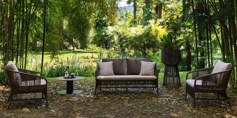 Varaschin Cricket Lounge Gartenmöbel in Braun, bestehend aus Sofa, Sesseln und Tisch, auf einer Terrasse mit üppigem Garten im Hintergrund.