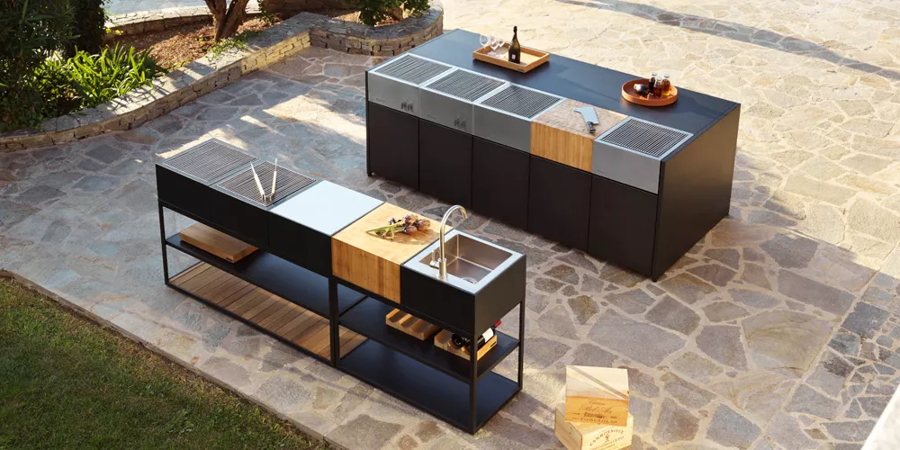 Roshults Kitchen Island in Schwarz und Holzoptik auf einer Terrasse, mit Grill und Spüle, umgeben von Pflanzen und Steinboden.
